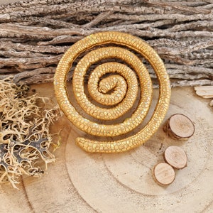 May include: A circular, coiled object with a textured, metallic gold surface featuring a repeating scale-like pattern. The form resembles a spiral or a coiled snake, with a smooth, rounded profile and a consistent golden finish throughout the entire piece.