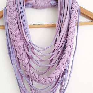 May include: A handmade lavender and light purple infinity scarf. The scarf features a braided design on each side and multiple strands of fabric hanging down. The scarf is displayed on a wooden hanger against a white background.