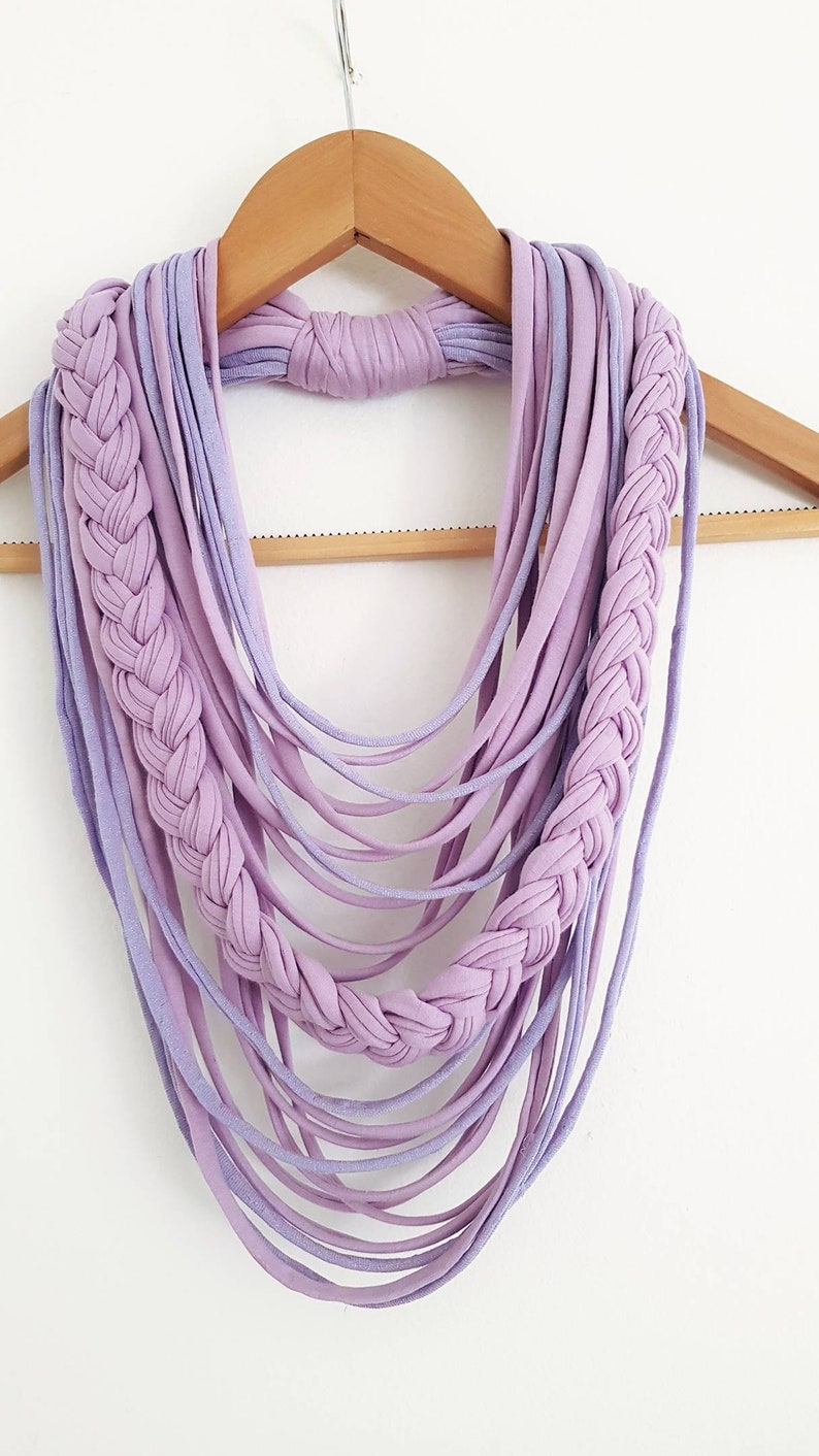 May include: A handmade lavender and light purple infinity scarf. The scarf features a braided design on each side and multiple strands of fabric hanging down. The scarf is displayed on a wooden hanger against a white background.