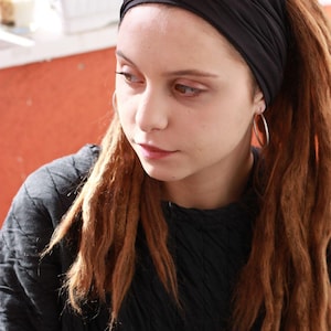May include: A person wearing a black headband and a black top. The person has long, reddish-brown dreadlocks and silver hoop earrings. The background is a blurred orange wall and a white window.