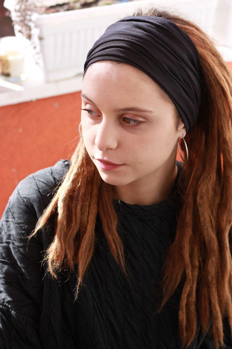 May include: A person wearing a black headband and a black top. The person has long, reddish-brown dreadlocks and silver hoop earrings. The background is a blurred orange wall and a white window.