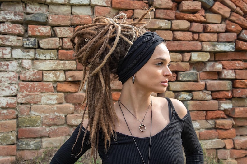 May include: A black fabric headband featuring a braided design worn with long brown dreadlocks. The accessory is paired with a black long-sleeve top, a silver hoop earring, and a layered necklace with a circular pendant against a brick wall.