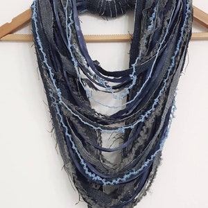 May include: A multi-strand infinity scarf constructed from various strips of denim, frayed fabric, and ribbon in shades of dark blue, charcoal gray, and light blue. The layered design features raw edges and a textured, distressed appearance.
