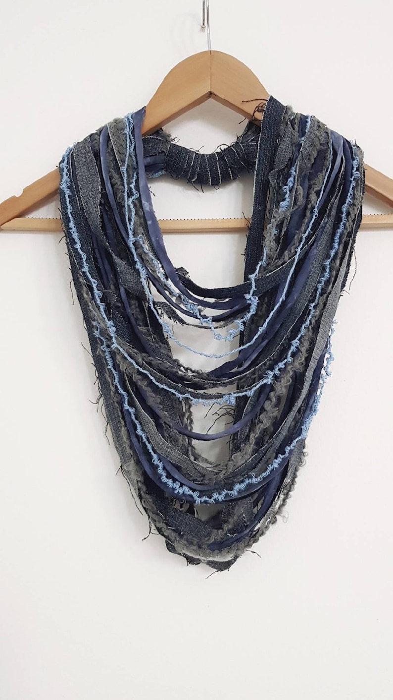 May include: A multi-strand infinity scarf constructed from various strips of denim, frayed fabric, and ribbon in shades of dark blue, charcoal gray, and light blue. The layered design features raw edges and a textured, distressed appearance.