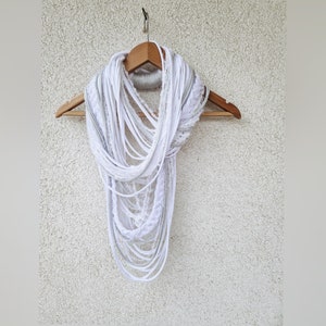 Puede incluir: Un pañuelo infinito blanco con un diseño en capas, exhibido en una percha de madera. El pañuelo presenta varias texturas, incluyendo secciones trenzadas y con flecos, creando un accesorio único y elegante. El pañuelo está hecho de un material suave y ligero.