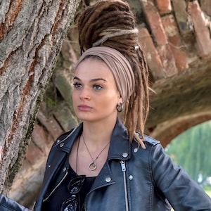 May include: A person wearing a black leather jacket, black top, and a tan head wrap. The person has dreadlocks and is wearing a necklace and sunglasses. The background includes a tree trunk and a brick structure.