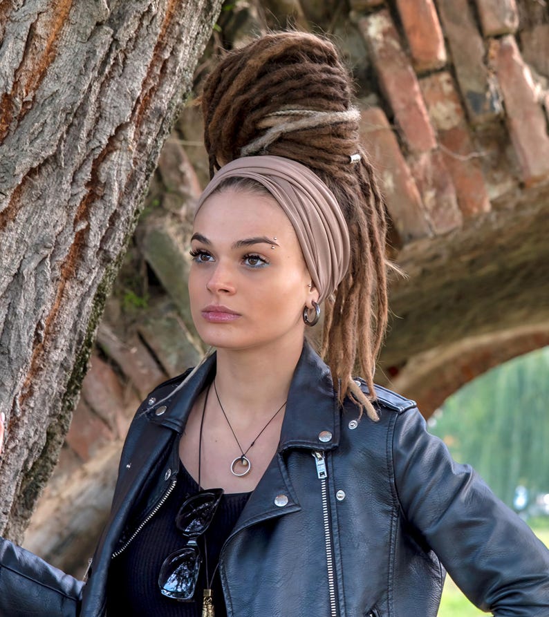 May include: A person wearing a black leather jacket, black top, and a tan head wrap. The person has dreadlocks and is wearing a necklace and sunglasses. The background includes a tree trunk and a brick structure.