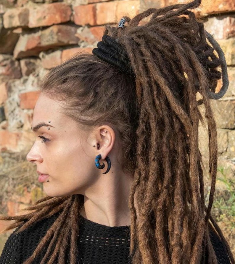 K&ouml;nnte beinhalten: Ein dunkelblauer und schwarzer spiralf&ouml;rmiger Ohrschmuck, der im Ohrl&auml;ppchen getragen wird. Der Ohrring hat ein geschwungenes, spitz zulaufendes Design mit glatter Oberfl&auml;che, das einen Kontrast zu den braunen Dreadlocks und dem schwarzen Oberteil bildet.