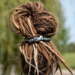 May include: A flexible, fabric-covered hair tie with a teal, black, and white floral pattern wrapped around a large bun of brown dreadlocks. The hair accessory is knotted at the center, securing the thick, textured locks in a high updo style.