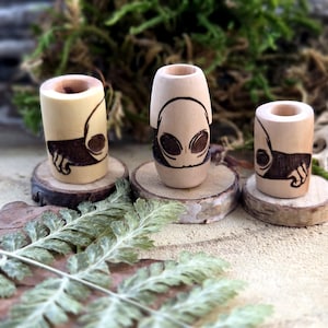 Peut inclure: Trois perles en bois avec des visages d'extraterrestres gravés dessus. Les perles sont sur de petits ronds en bois.