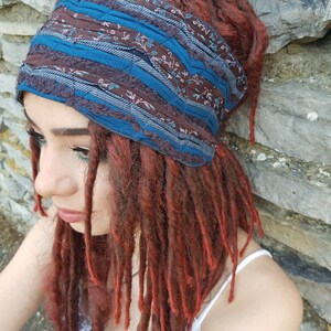 May include: A wide fabric headband featuring a patchwork design of dark brown floral fabric, solid teal blue strips, and textured dark blue fabric with a ribbed pattern. The headband is worn around the head, contrasting with long, auburn-colored dreadlocks.