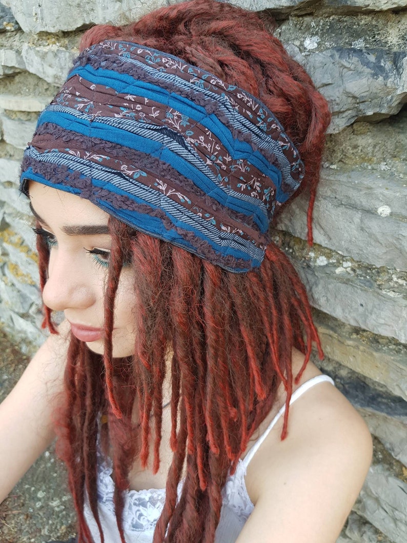 May include: A wide fabric headband featuring a patchwork design of dark brown floral fabric, solid teal blue strips, and textured dark blue fabric with a ribbed pattern. The headband is worn around the head, contrasting with long, auburn-colored dreadlocks.