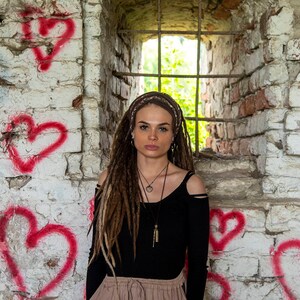 May include: A person with long brown dreadlocks wearing a black long-sleeve shirt with shoulder cutouts and a tan skirt. The person has a layered necklace and a brown headband, positioned in front of a white brick wall painted with red hearts.