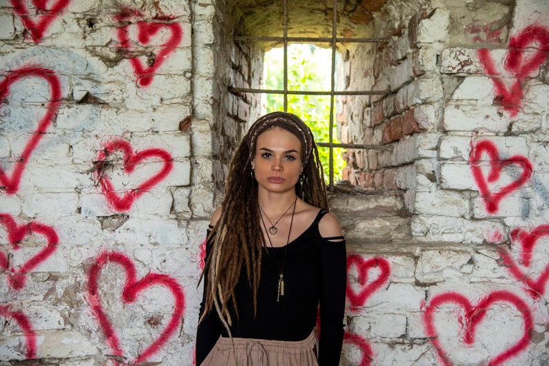 May include: A person with long brown dreadlocks wearing a black long-sleeve shirt with shoulder cutouts and a tan skirt. The person has a layered necklace and a brown headband, positioned in front of a white brick wall painted with red hearts.