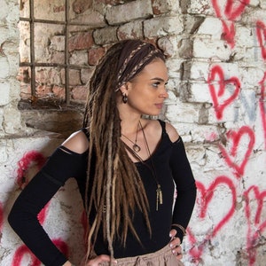 A woman with long brown dreadlocks wearing a black long-sleeved shirt with cut-out shoulders, a brown headband, and multiple necklaces. She is standing in front of a white brick wall with red hearts painted on it.