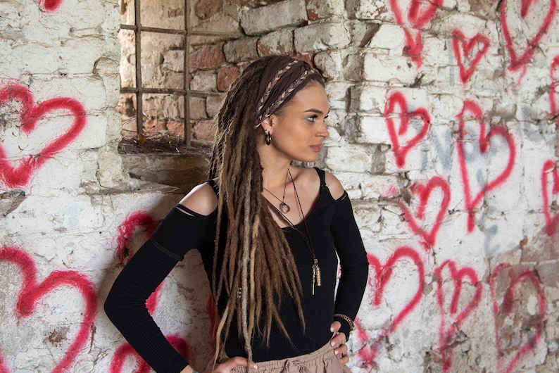A woman with long brown dreadlocks wearing a black long-sleeved shirt with cut-out shoulders, a brown headband, and multiple necklaces. She is standing in front of a white brick wall with red hearts painted on it.