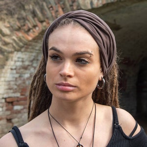 Pode incluir: Uma mulher com dreadlocks usa uma faixa de cabeça marrom e um brinco de argola prateado. Ela tem um piercing no nariz e um piercing na sobrancelha prateados. Ela está vestindo um top preto sem mangas com um colar prateado.