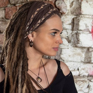 May include: A dark brown fabric headband featuring a braided detail across the top. The headband is worn over long brown dreadlocks. A silver-toned ring pendant hangs from a thin black cord necklace, and a small silver hoop earring is visible.