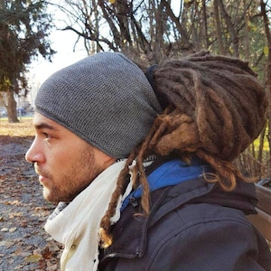 Peut inclure: Un homme avec des dreadlocks porte un bonnet en tricot gris, une écharpe blanche et une veste noire. Il est debout à l'extérieur devant un arbre.