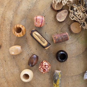 May include: A selection of handcrafted wooden beads and small wood slices in various shapes and colours. The beads feature cylindrical, oval, and spherical designs, with natural wood grain and painted patterns. The arrangement is on a light brown, textured surface.