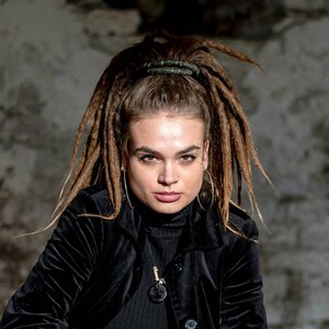 K&ouml;nnte beinhalten: Eine Person mit langen braunen Dreadlocks, die zu einem hohen Pferdeschwanz gebunden sind, tr&auml;gt eine schwarze Samtjacke &uuml;ber einem schwarzen gerippten Rollkragenpullover. Die Person tr&auml;gt gro&szlig;e silberne Creolen, ein Augenbrauenpiercing und einen Anh&auml;nger.
