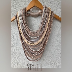 Op de afbeelding: Een meerstrengs ketting in beige, bruin en cr&egrave;metinten, tentoongesteld op een houten hanger. De ketting heeft verschillende texturen, waaronder gevlochten touw, gladde koorden en gefranjerde elementen. De tekst "STYLE 1" staat onderaan.