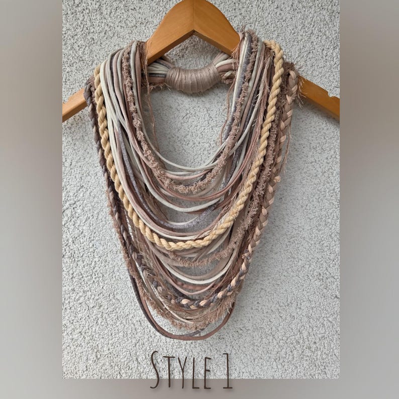 Op de afbeelding: Een meerstrengs ketting in beige, bruin en cr&egrave;metinten, tentoongesteld op een houten hanger. De ketting heeft verschillende texturen, waaronder gevlochten touw, gladde koorden en gefranjerde elementen. De tekst "STYLE 1" staat onderaan.