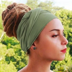 May include: An olive green turban-style headband. The fabric is soft and wraps around the head, with a knot detail at the front. The background is a blurred view of green foliage.