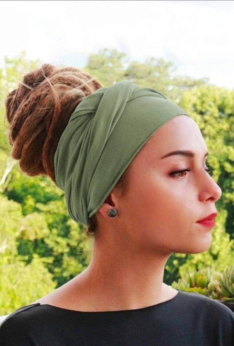 May include: An olive green turban-style headband. The fabric is soft and wraps around the head, with a knot detail at the front. The background is a blurred view of green foliage.