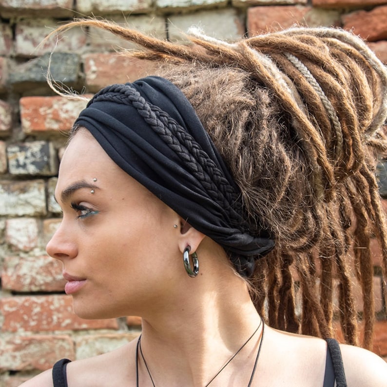 May include: A black fabric headband featuring a braided detail across the top. The headband is worn over long brown dreadlocks. The accessory is made of a soft, flexible material that wraps around the head, providing a secure fit for hair.