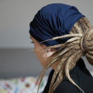 K&ouml;nnte beinhalten: Ein marineblaues Kopftuch bedeckt den Kopf, mit langen, hellbraunen Dreadlocks, die zu einem Dutt frisiert sind. Die Person tr&auml;gt ein schwarzes Oberteil. Der Hintergrund ist ein neutrales Grau.