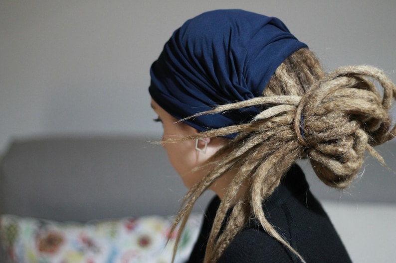 K&ouml;nnte beinhalten: Ein marineblaues Kopftuch bedeckt den Kopf, mit langen, hellbraunen Dreadlocks, die zu einem Dutt frisiert sind. Die Person tr&auml;gt ein schwarzes Oberteil. Der Hintergrund ist ein neutrales Grau.