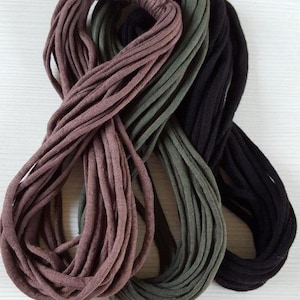 May include: Three circular bundles of fabric yarn in solid colors of mauve, olive green, and black. Each bundle consists of multiple thin, continuous strands of jersey-like material looped together to form a long, flexible, and soft corded structure.