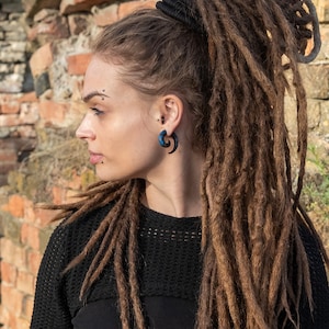 A woman with long brown dreadlocks pulled back in a ponytail with a black hair tie. She is wearing a black sweater and has a blue and black spiral ear gauge in her right ear.