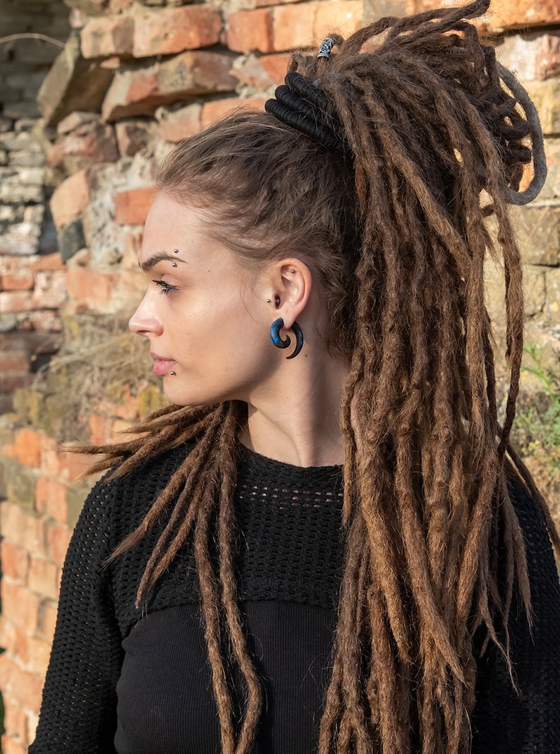 A woman with long brown dreadlocks pulled back in a ponytail with a black hair tie. She is wearing a black sweater and has a blue and black spiral ear gauge in her right ear.