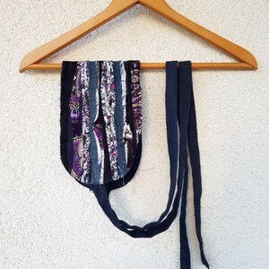 May include: A colorful fabric strap with a blue fabric tie hanging on a wooden hanger. The strap is made of various fabrics, including denim, and has a purple and white paisley pattern.