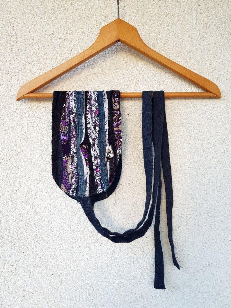 May include: A colorful fabric strap with a blue fabric tie hanging on a wooden hanger. The strap is made of various fabrics, including denim, and has a purple and white paisley pattern.