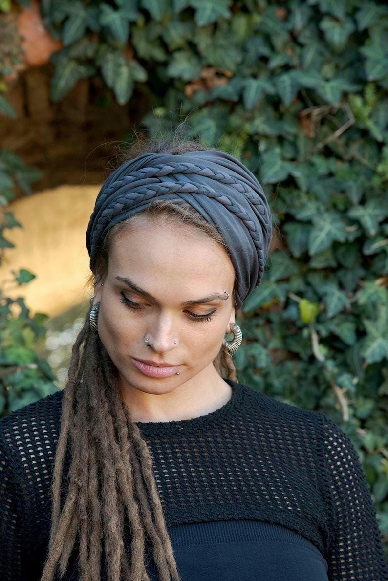 May include: A dark gray fabric headband featuring a wide, gathered design with multiple braided strands across the top. The headband is constructed from a soft, matte material that wraps around the head, creating a layered and textured appearance.