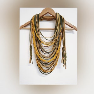 Puede incluir: Un collar largo y de múltiples capas hecho de tiras de tela en tonos de amarillo mostaza y verde bosque. El collar está atado con un nudo en la parte inferior y tiene bordes deshilachados.