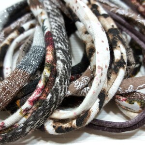 May include: A collection of thin, circular fabric headbands featuring a variety of patterns and textures. The assortment includes brown, black, white, and tan colors with speckled, animal print, and solid knit designs in a layered, coiled arrangement.