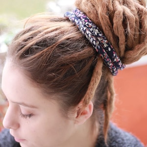 Pu&ograve; includere: Un elastico per capelli in tessuto blu scuro con un motivo floreale multicolore avvolto attorno a un grande chignon di dreadlocks marrone chiaro. L'accessorio per capelli &egrave; posizionato saldamente alla base dello chignon, contrastando i capelli.