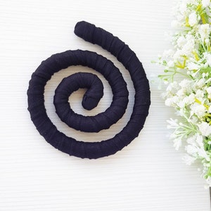 May include: A black fabric-covered flexible cord coiled into a spiral shape. The cord features a uniform, tightly wrapped textile texture throughout its entire length, creating a continuous circular form that rests flat against the surface.