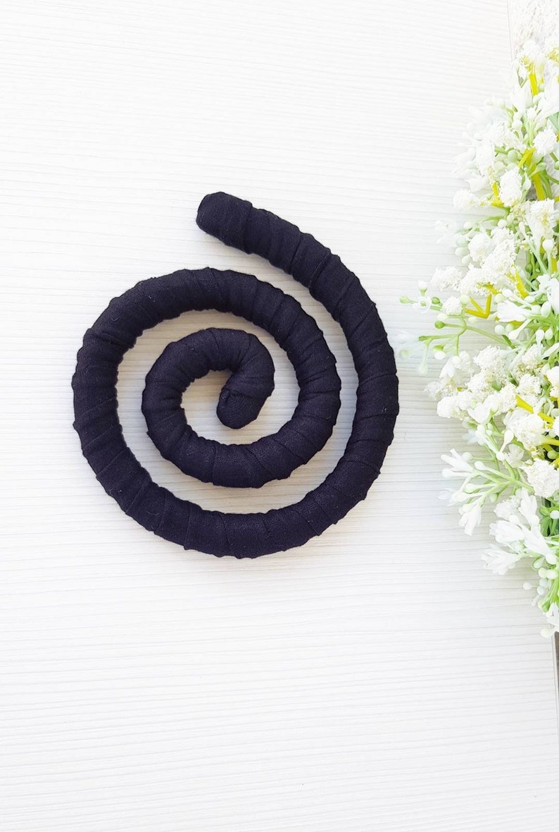 May include: A black fabric-covered flexible cord coiled into a spiral shape. The cord features a uniform, tightly wrapped textile texture throughout its entire length, creating a continuous circular form that rests flat against the surface.