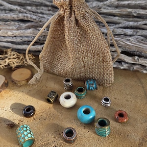 Könnte beinhalten: Eine Sammlung von Perlen in verschiedenen Farben und Designs, darunter Türkis, Weiß und Metallic-Oberflächen, liegt auf einer Holzoberfläche. Ein kleiner Jute-Kordelzugbeutel befindet sich im Hintergrund und deutet auf die Aufbewahrung oder Verpackung der Perlen hin.