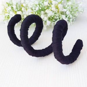 May include: A black fabric-wrapped spiral accessory featuring a continuous coiled design. The material is tightly wound around the structure, creating a textured surface. The object is shaped like a spring with multiple loops, presenting a uniform dark color throughout its entire length.