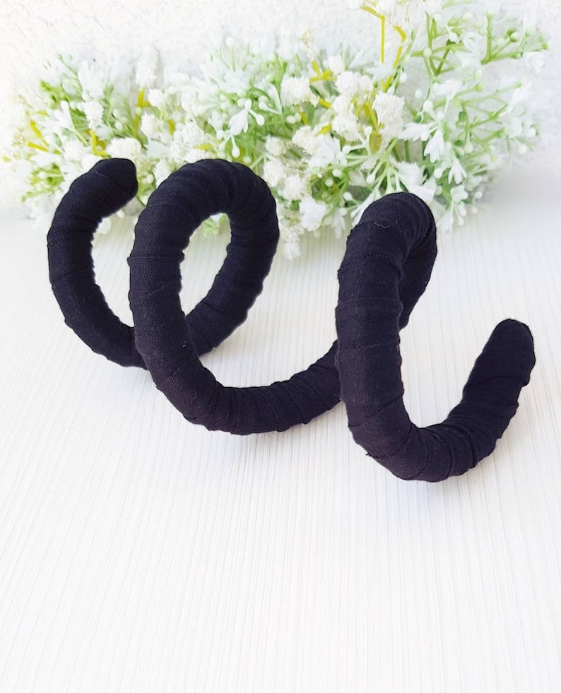 May include: A black fabric-wrapped spiral accessory featuring a continuous coiled design. The material is tightly wound around the structure, creating a textured surface. The object is shaped like a spring with multiple loops, presenting a uniform dark color throughout its entire length.