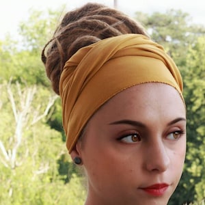 May include: Mustard yellow head wrap. The fabric is wrapped around the head and tied at the back. The background is a blurred view of green foliage. The person in the image has brown dreadlocks and is wearing a black top.