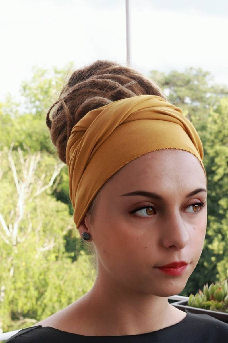 May include: Mustard yellow head wrap. The fabric is wrapped around the head and tied at the back. The background is a blurred view of green foliage. The person in the image has brown dreadlocks and is wearing a black top.