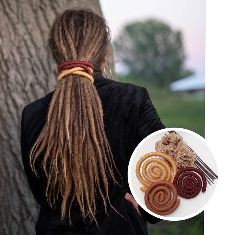 May include: A set of three flexible, spiral-shaped hair accessories in mustard yellow, brown, and deep red, accompanied by a cluster of desert rose crystal and thin dark sticks, shown alongside a person with long brown dreadlocks.