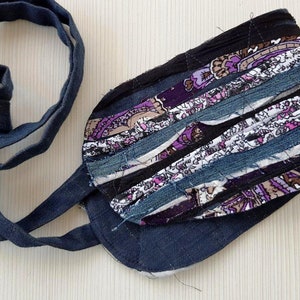 May include: A handmade fabric belt with a dark blue denim base and a colorful patchwork design featuring purple, pink, white, and brown fabric strips. The belt is tied with a long, dark blue fabric tie.